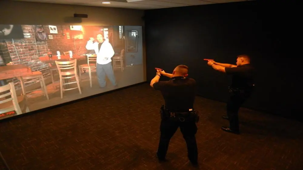 Two officers engage in firearm training simulation, confronting a projected suspect.