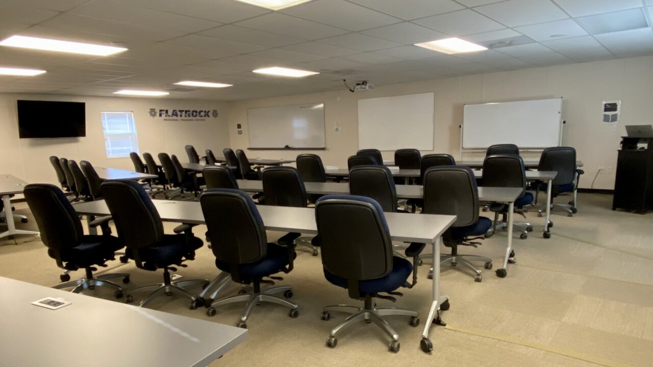 Classroom setup with rows of tables and chairs, whiteboards, and a screen, showcasing a learning environment.