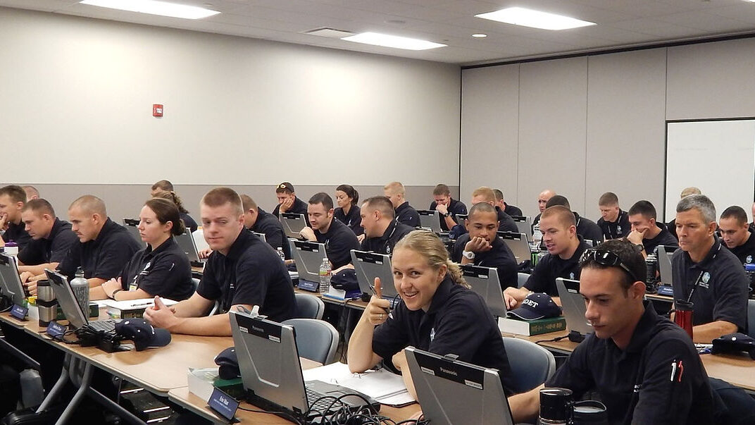 Classroom with uniformed individuals seated at desks working on laptops, one person smiling and gesturing thumbs up