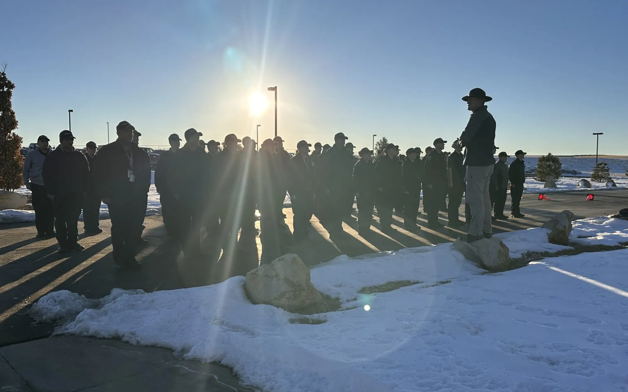 Group of people in hats standing in snow, listening to a speaker at sunrise