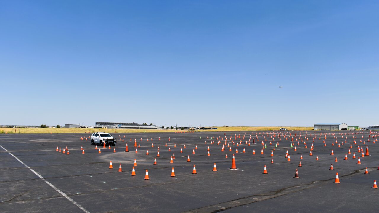 White SUV navigating through a large parking lot with scattered orange traffic cones under a clear blue sky.