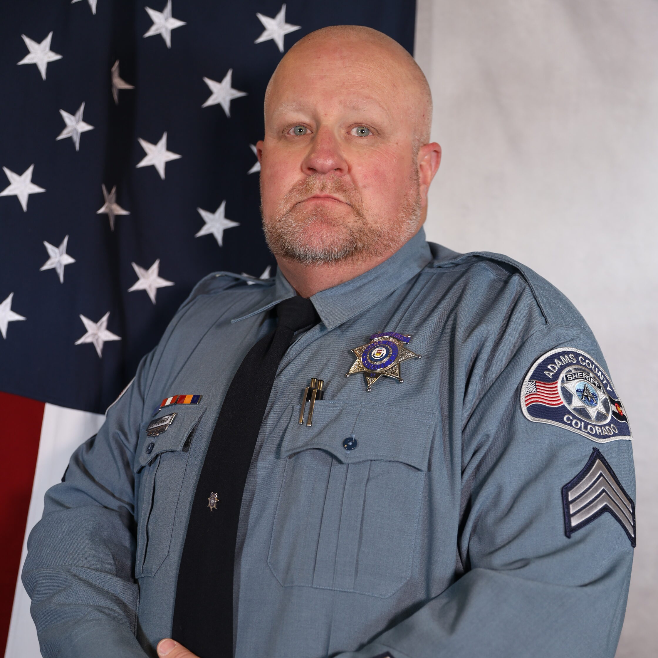 Dummar-Derek in police uniform, standing with hands clasped, U.S. flag background, Adams County badge visible.