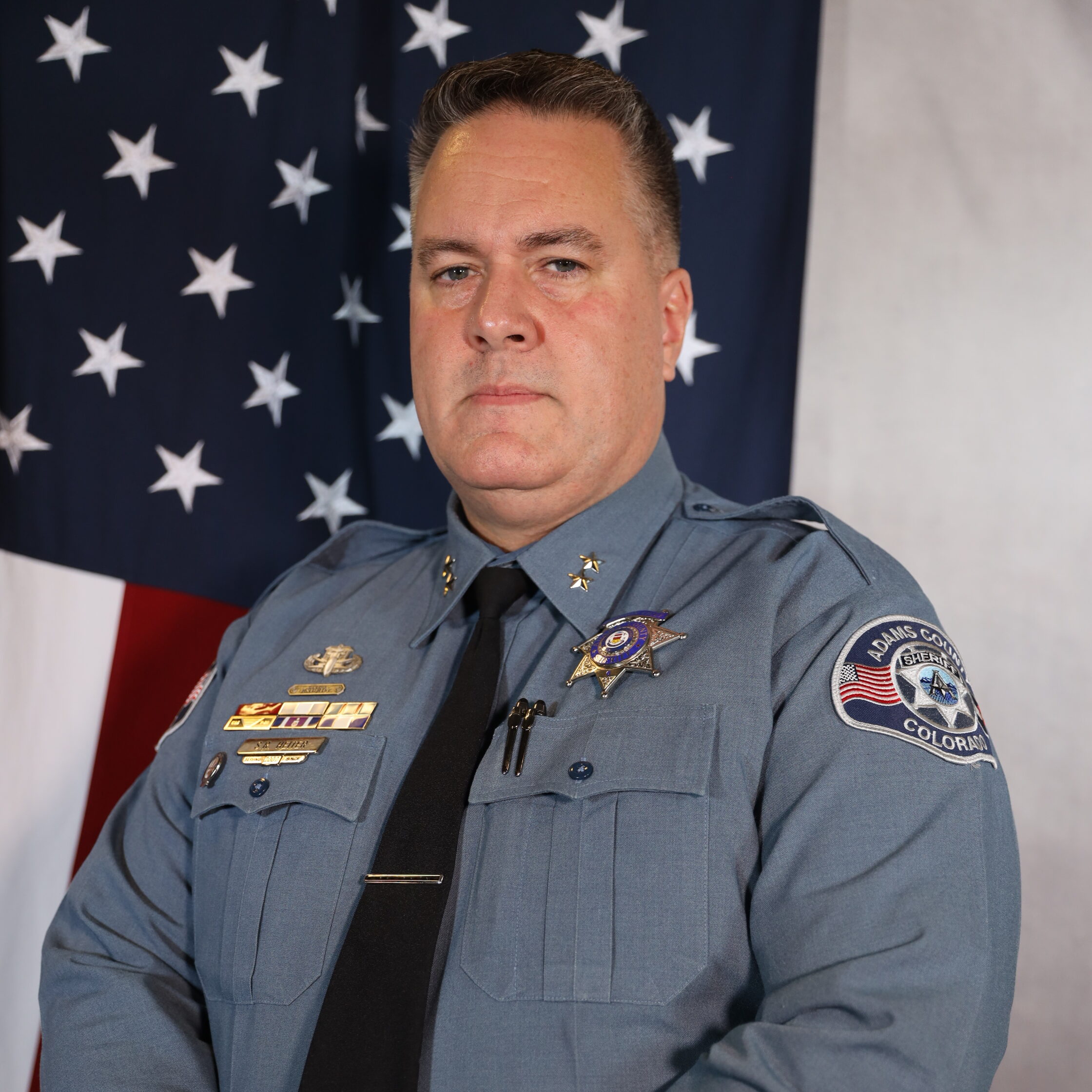 Heiter-Shane in uniform, standing with American flag backdrop, serious expression.