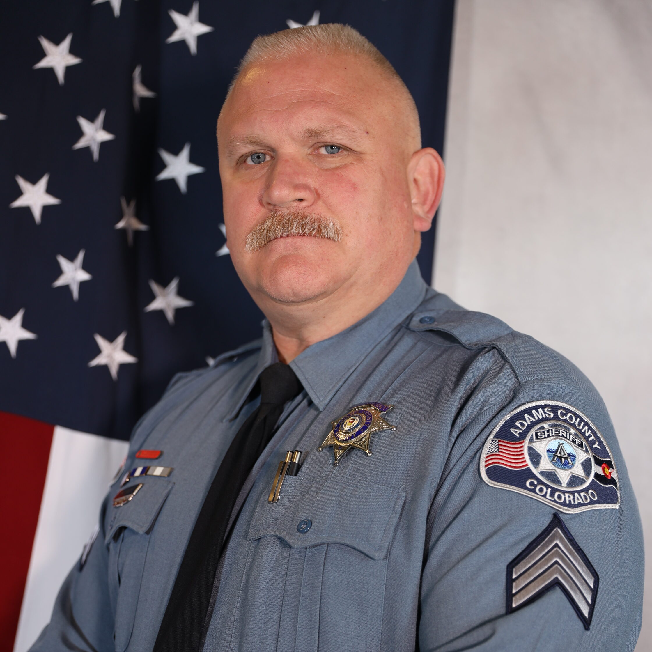 Moore-Gabriel.jpg: Uniformed officer poses confidently, U.S. flag behind. Adams County Sheriff Colorado patch visible.